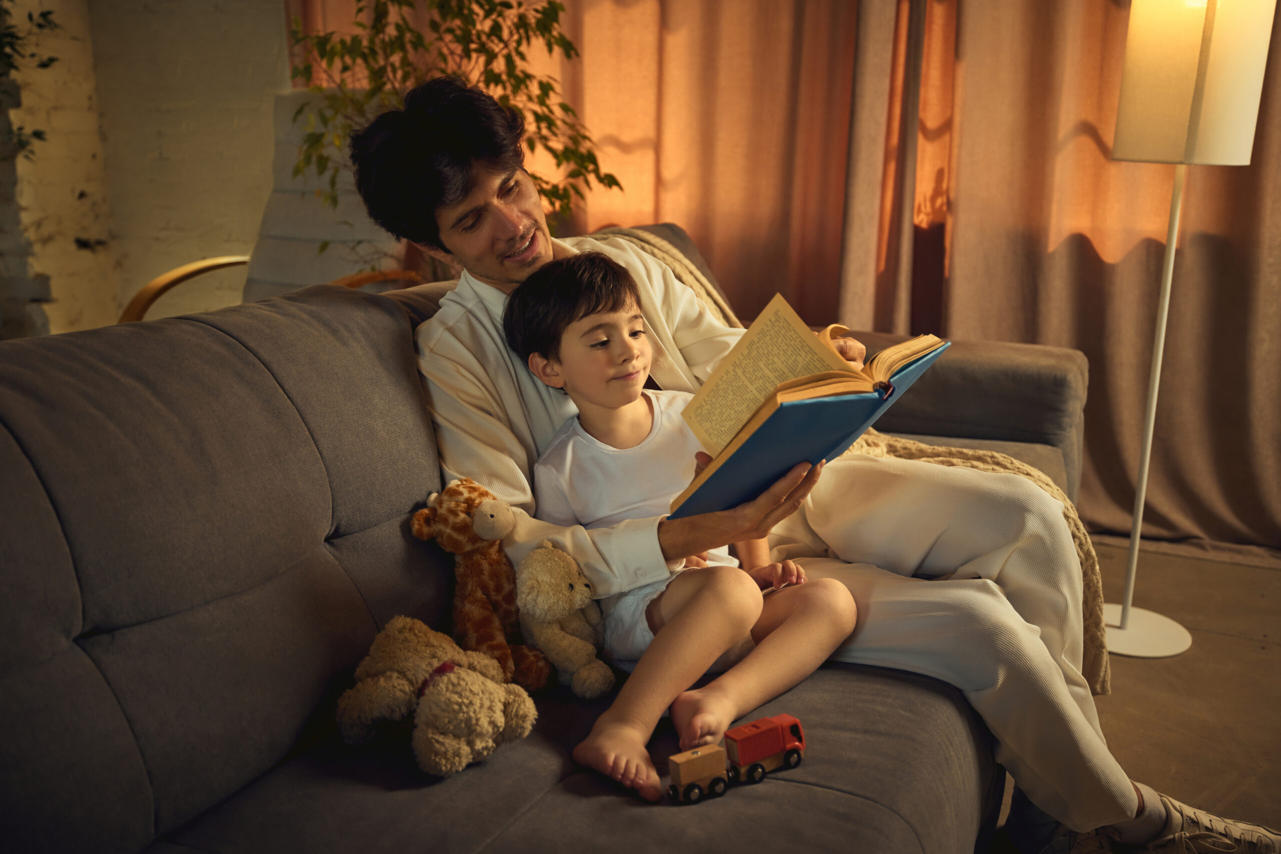A father engages his son with a bedtime story while they sit on a comfortable sofa amidst warm lighting and scattered toys. Concept of parent-child bond and early childhood education